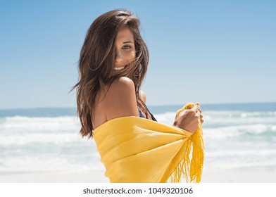 Cheerful young woman in yellow sarong at beach. Happy smiling girl enjoying the beach and looking at camera. Latin tanned woman feeling refreshed in yellow scarf during summer vacation.