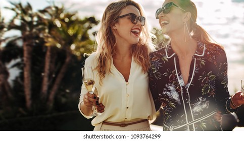 Cheerful young women walking together with glass of wine. Stylish female friends enjoying themselves outdoors.