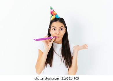 Cheerfully Surprised Young Woman In Festive Birthday Cap, Blowing Kazoo, Standing Against White Background