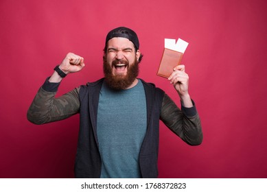 Cheering bearded man is holding passport with tickets while making winner gesture over red pink background.