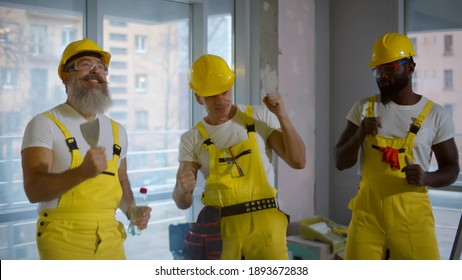 Cheering Diverse Construction Workers Dancing And Laughing In Renovating Apartment. Portrait Of Multiethnic Male Builders In Hardhat And Overall Having Fun And Dancing During Break