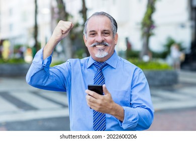 Cheering Latin American Senior Businessman Receiving Good News On Phone Outdoor In City In Summer