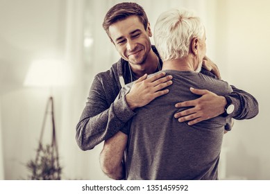 Cheering Up Sick Dad. Smiling Beaming Handsome Young Man In Grey Hoodie Embracing Grey-haired Dad In Years Cheering Him Up