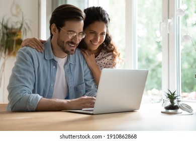 Cheering Spouses Having Warm Relations Hug By Desk At Home Make Shopping Purchases Online At Web Store. Friendly Young Couple In Love Look At Computer Screen Planning Wedding Choosing Tour On Vacation