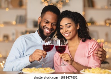 Cheers. Beautiful lovely young african american couple sitting at table in restaurant or at home, drinking red wine, holding glasses during romantic dinner. Smiling black man embracing his happy woman