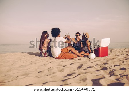 Cheers to friends! Cheerful young people spending nice time together while sitting on the beach and drinking beer