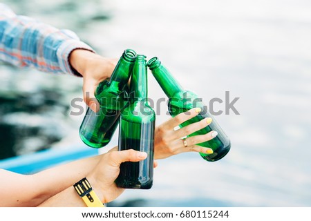 Cheers! Group of young people holding and toasting with green bottles of beer on river background. Celebrating oktoberfest with friends outdoor. Barbecue, hanging out, relaxing concepts.