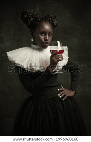 Cheers. Medieval African young woman in black vintage dress with big white collar isolated on dark green background. Concept of comparison of eras, modernity and renaissance. White pearls