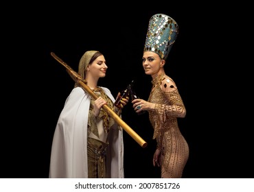 Cheers. Portrait Of Young Woman In Image Of Nefertiti In Art Performance With With Her Servant Drinking Beer Isolated On Dark Background. Model Like Famous Queen Of Egypt. Comparison Of Eras Concept.