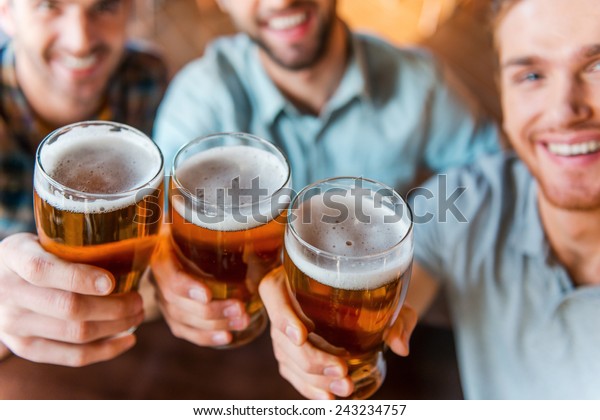 Cheers to success! Top view of three happy young men in casual wear toasting with beer while sitting in bar together