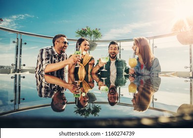 Cheers. Two Young Couples Spending Time At Rooftop Bar With Alcohol Drinks. They Are Toasting And Laughing