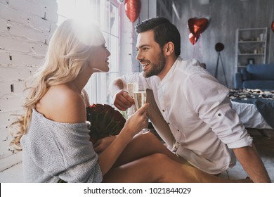 Cheers to us! Beautiful young couple drinking champagne and smiling while sitting in the bedroom - Shutterstock ID 1021844029