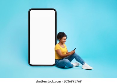 Fröhliche Afrikanisch-amerikanische Dame sitzend in der Nähe des großen Mobiltelefons mit leerem weißem Bildschirm, mit dem mobilen Gerät, Überprüfung der neuen coolen App auf blauem Studiohintergrund, Muster für Website- oder Anwendungsdesign