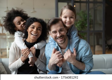 Cheery African and Caucasian girls cuddle piggy back multi-racial parents loving mom and dad seated on sofa in living room. Happy multinational family portrait, new home funny weekend together concept - Shutterstock ID 1756081292