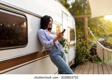 Cheery Asian lady leaning on motorhome, using cellphone on autumn camping trip in countryside. Funky young woman with mobile device browsing web, chatting online, watching video outdoors