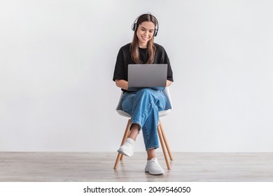 Cheery Caucasian woman in headphones having online video call on laptop, sitting on chair against white wall, full length. Millennial lady participating in webinar, communicating remotely on internet - Shutterstock ID 2049546050