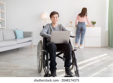 Cheery disabled teen boy in wheelchair with laptop smiling at camera indoors, his mother making cleanup on background. Happy impaired youth studying remotely from home, using portable computer