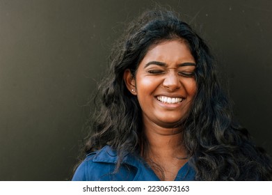 Cheery indian woman laughing with eyes closed while standing over dark background outdoors - Shutterstock ID 2230673163