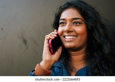 Cheery Indian Woman Talking On Cellphone While Standing Over Dark Background Outdoors