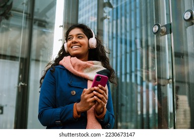 Cheery indian woman using mobile phone and wireless headphones while walking through city street outdoors - Shutterstock ID 2230673147