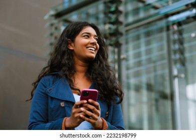 Cheery indian woman using mobile phone while walking through city street - Shutterstock ID 2232198799