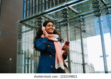 Cheery indian woman using mobile phone and wireless headphones while walking through city street outdoors - Shutterstock ID 2232198821