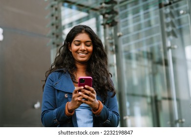Cheery indian woman using mobile phone while walking through city street - Shutterstock ID 2232198847