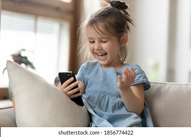 Cheery Little Girl Watching Cartoons On Smart Phone. Kid Girl Sit On Couch Hold Gadget Spend Time On Internet Fun Website For Children. New Generation And Modern Tech Overuse, Parental Control Concept