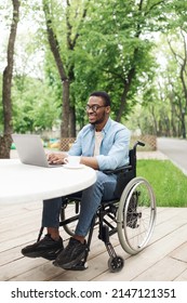 Cheery Millennial Black Man In Wheelchair Using Laptop For Online Work At Outdoor Cafe, Having Business Meeting On Web. Smiling African American Guy With Disability Working On Remote Project At Park