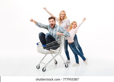 Cheery Mother And Her Daughter Giving A Ride On A Shopping Trolley To Their Father Isolated Over White Background