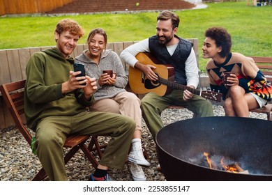 Cheery positive multinational friends drinking hot tea and using smartphone while resting around campfire with guitar - Shutterstock ID 2253855147