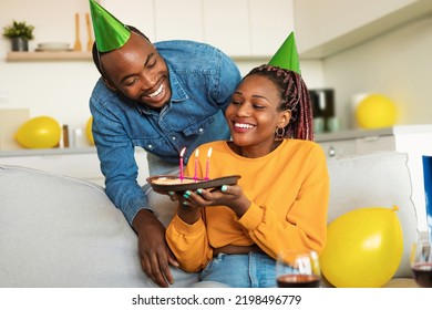 Cheery Young Black Couple In Party Hats Celebrating Birthday, Woman Holding Birthday Pie With Lit Candles. African American Family Enjoying Domestic B-day Party