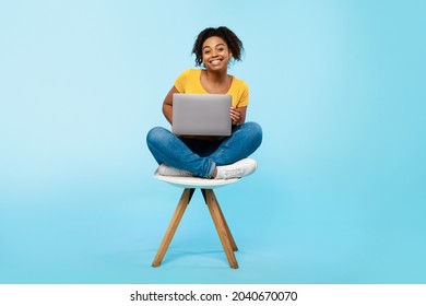 Cheery young black woman working online, sitting on chair and using laptop on blue studio background, full length. Cheerful African American lady surfing internet on portable pc - Shutterstock ID 2040670070