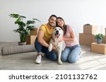 Cheery young international couple with cute golden retriever dog sitting on floor of new home on relocation day. Happy millennial husband and wife with cute pet moving to new house