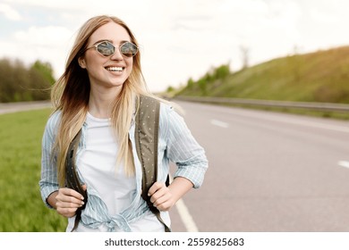 Cheery young lady in casual summer clothes walking along road in countryside, hitchhiking for ride outdoors. Beautiful millennial blonde woman with backpack traveling by autostop - Powered by Shutterstock