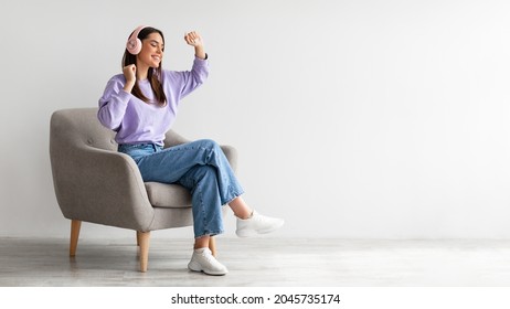 Cheery young lady in headphones listeing to music and dancing while sitting in armchair against white studio wall, banner design with free space. Carefree Caucasian woman moving to favorite song - Shutterstock ID 2045735174