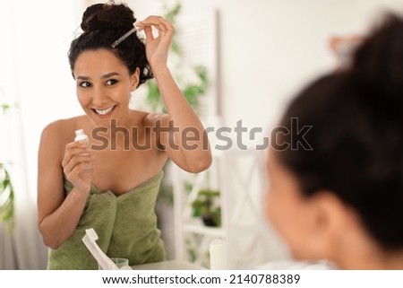 Cheery young woman in towel applying serum or natural oil on her scalp near mirror at home, copy space. Organic cosmetics for haircare, damaged dry hair treatment, daily beauty routine concept