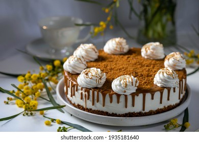 Cheese Cake Decorated With Whipped Cream, Caramel And Cookie Crumbs, Which Stands On A White Plate With Mimosa Flowers