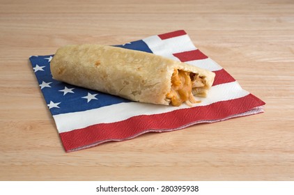 A Cheese And Chicken Chimichanga That Has Been Bitten On A Flag Motif Napkin Atop A Wood Table Top.