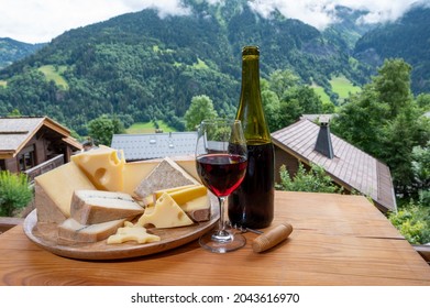 Cheese collection, wooden board with French cheeses comte, beaufort, abondance, emmental, morbier, red wine from Savoie and french mountains village in Haute-Savoie in summer on background