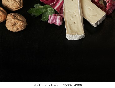 Cheese delikatessen closeup on black stone desk surface. Camembert or brie circle in brown kraft paper decorated with basil and pieces of cherry tomatoes, top view image with copy space.
