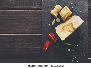 Cheese delikatessen closeup on black stone desk at wooden surface background. Parmesan pieces decorated with basil and cherry tomatoes, top view image with copy space, soft toning