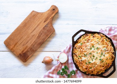 Cheese Noodles In Iron Cast Pan And Empty Cutting Board On A White Table. Above View Of German Traditional Cheese Spaetzle With Bacon. Swabian Food.