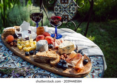 Cheese Platter With Different Cow And Goat Cheeses, Cured Meat, Nuts, Summer Fruits And Berries, Jam And Rye Bread On Wooden Board With Red Wine On Little Rustic Table In The Garden.