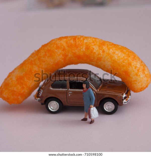Cheese Puff on a tiny car with woman holding\
grocery bags. Giant cheesy. Concept of food delivery or ordering\
online. Transportation of food can be overwhelming. Studio scene\
using miniature people.