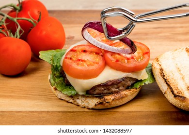 Cheeseburger With Havarti Cheese Tomatoes And Red Onions And Lettuce On A Toasted Cornmeal Bun On A Wooden Table With Bun Open Showing Onion Being Placed On The Burger