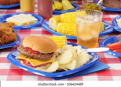 A Cheeseburger With Potato Chips And Corn On The Cob On A Picnic Table Loaded With Food