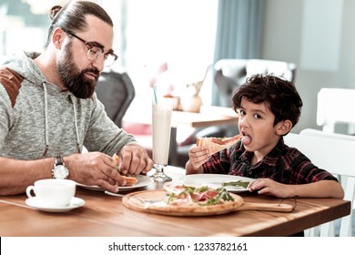 Cheesy pizza. Cute dark-haired son eating yummy cheesy pizza with his father while having lunch in cafeteria