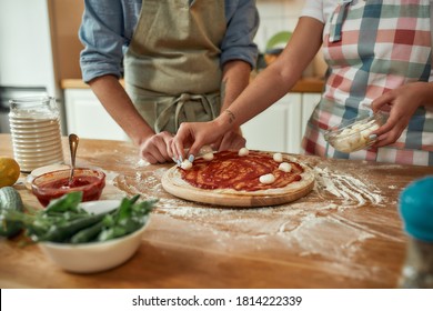 For Cheesy Taste. Cropped Shot Of Couple Making Pizza Together At Home. Man In Apron Adding, Applying Tomato Sauce On The Dough While Woman Adding Mozzarella Cheese. Selective Focus. Horizontal Shot