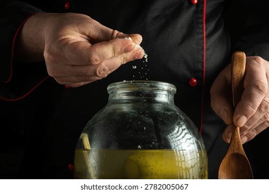 A chef is adding salt to a jar of pickles in a kitchen. The chef focuses on the jar while holding a wooden spoon. Light shines through the jar, showing ingredients inside.